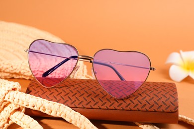 Stylish heart shaped sunglasses and brown leather case on pale orange background