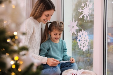 Happy mother and daughter near window decorated with paper snowflakes indoors