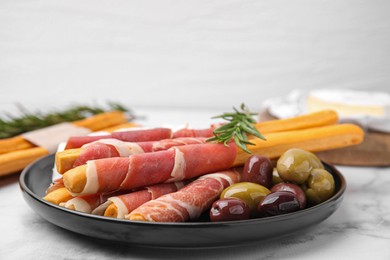 Photo of Plate of delicious grissini sticks with prosciutto and olives on white marble table, closeup