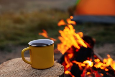 Yellow mug with hot drink on wooden stump near bonfire outdoors, space for text. Camping season