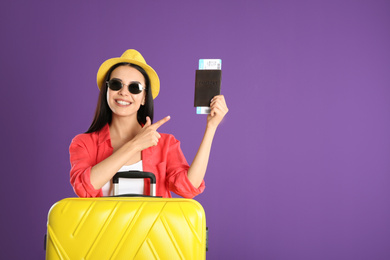 Beautiful woman with suitcase and ticket in passport for summer trip on purple background. Vacation travel