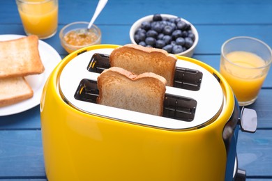 Yellow toaster with roasted bread, glasses of juice, blueberries and jam on blue wooden table, closeup