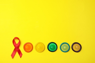Flat lay composition with colorful condoms and red ribbon on yellow background, space for text. LGBT concept