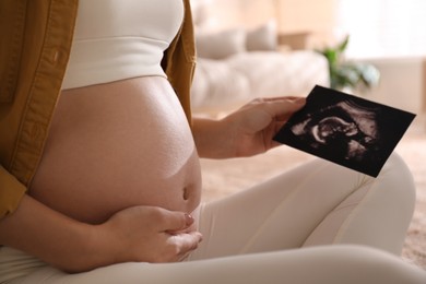 Pregnant young woman holding ultrasound picture near her belly at home, closeup