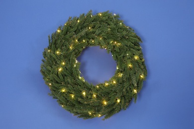 Christmas wreath made of fir branches with string lights on blue background, top view