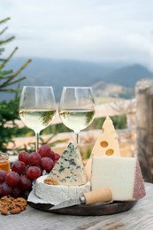 Different types of delicious cheeses, snacks and wine on wooden table against mountain landscape