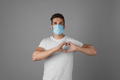 Man in protective mask making heart with hands on grey background