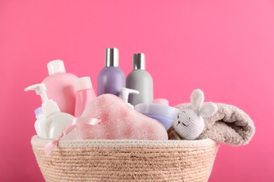 Different baby cosmetic products, bathing accessories and toy in wicker basket on pink background, closeup