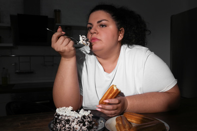 Depressed overweight woman eating sweets in kitchen at night