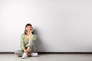 Beautiful young girl sitting on floor near white wall. Space for text