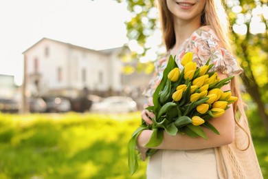 Teenage girl with bouquet of yellow tulips in city park on sunny day, closeup. Space for text