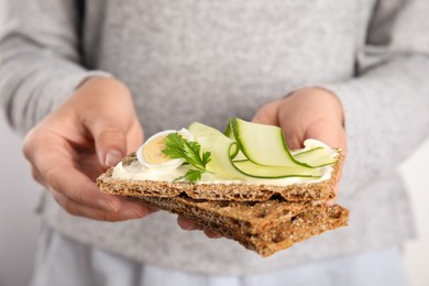 Woman holding fresh rye crispbreads with quail egg, cream cheese and cucumber slices on light background, closeup