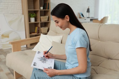 Young woman coloring antistress page on sofa in living room