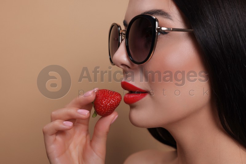 Attractive woman in fashionable sunglasses holding strawberry against beige background, closeup. Space for text