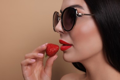 Attractive woman in fashionable sunglasses holding strawberry against beige background, closeup. Space for text