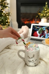 Photo of MYKOLAIV, UKRAINE - DECEMBER 23, 2020: Woman with sweet drink watching The Grinch movie on laptop near fireplace at home, closeup. Cozy winter holidays atmosphere
