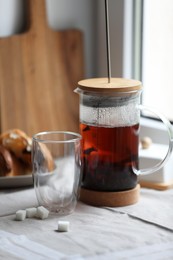 Teapot with freshly brewed tea, empty glass and sugar cubes on table near window