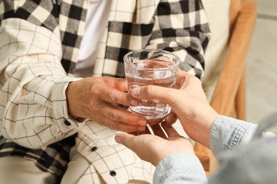 Caretaker giving glass of water to elderly woman indoors, closeup
