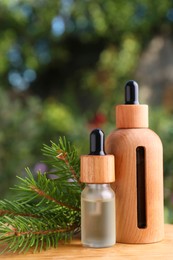 Bottles of pine essential oil and branches on wooden table against blurred background, closeup