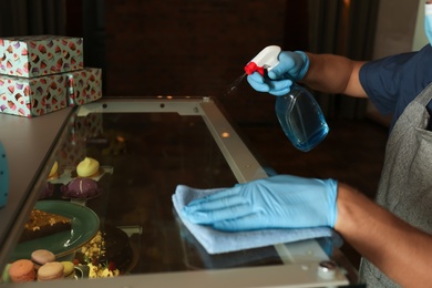 Worker in gloves disinfecting dessert showcase at cafe, closeup