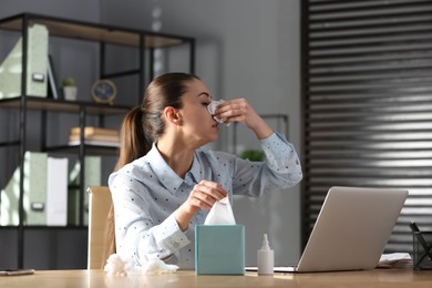 Ill businesswoman at table with nasal spray and box of paper tissues in office
