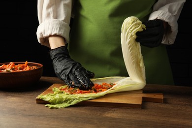Photo of Woman preparing spicy cabbage kimchi at wooden table against dark background, closeup