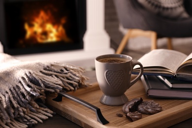 Cup of coffee, broken cookie and books on wooden table near fireplace indoors. Cozy atmosphere