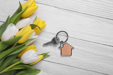 Photo of Beautiful spring flowers and key with trinket in shape of house on white wooden table, flat lay
