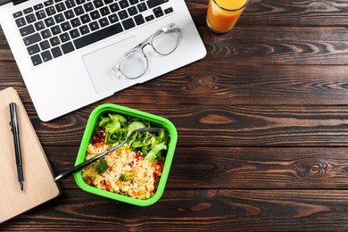 Photo of Container of tasty food, fork, laptop, glass of juice, and notebook on wooden table, flat lay with space for text. Business lunch