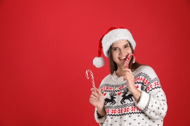 Pretty woman in Santa hat and Christmas sweater holding candy canes on red background, space for text