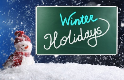 Text Winter Holidays on school chalkboard against decorative snowman in snowdrift