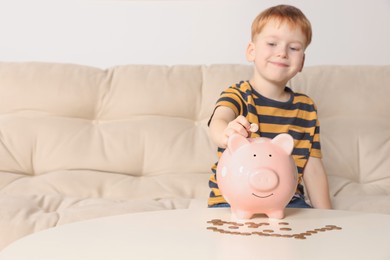 Cute little boy putting coin into piggy bank at table indoors, focus on hand. Space for text