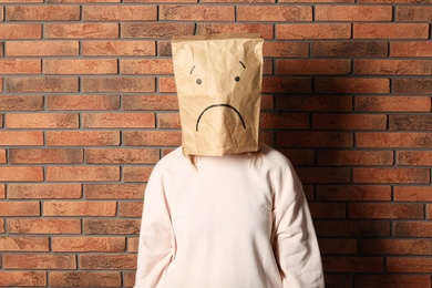 Woman wearing paper bag with drawn sad face near brick wall