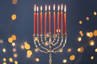 Silver menorah with burning candles against blue background and blurred festive lights. Hanukkah celebration