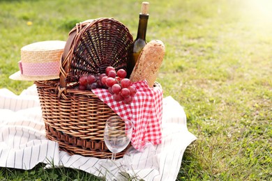Wicker basket with bottle of wine, bread, grapes, straw hat, napkin, glass and blanket on green grass outdoors, space for text. Picnic season