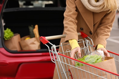 Photo of Young woman with shopping cart full of groceries near her car outdoors, closeup