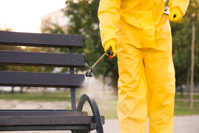 Person in hazmat suit disinfecting bench in park with sprayer, closeup. Surface treatment during coronavirus pandemic