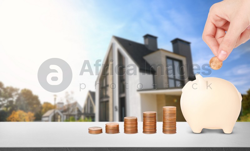 Woman putting coin into piggy bank at stone table and blurred view of beautiful house. Mortgage concept