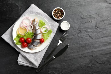 Photo of Sliced salted herring fillet served with lettuce, onion rings, cherry tomatoes and lemon on black table, flat lay. Space for text