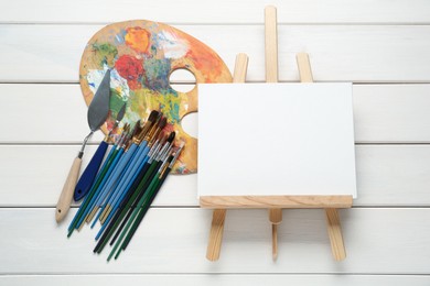 Easel with blank canvas, brushes and palette on white wooden table, flat lay. Space for text