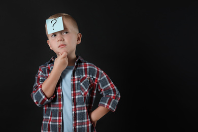 Pensive boy with question mark sticker on forehead against black background. Space for text