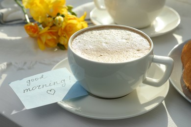 Cup of coffee, flowers and card with phrase GOOD MORNING! on white wooden table