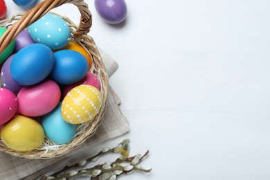 Colorful Easter eggs in wicker basket and willow branches on white wooden table, flat lay. Space for text