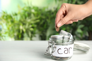 Woman putting coin into jar with tag CAR on white wooden table, closeup. Space for text