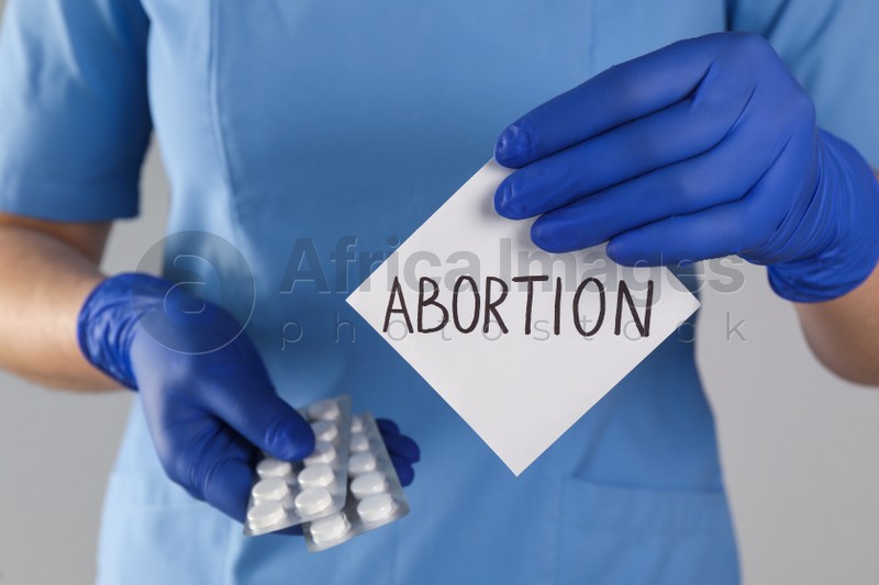 Doctor holding pills and paper card with word ABORTION on light grey background, closeup