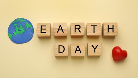 Phrase Earth Day made with wooden cubes, model of planet and red heart on beige, flat lay