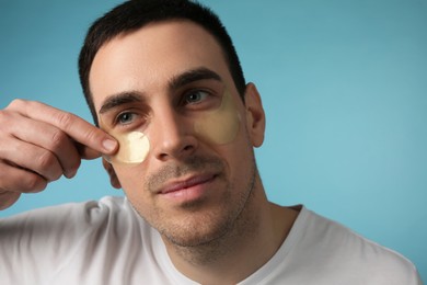 Young man applying under eye patches on light blue background
