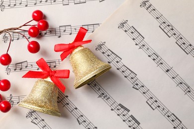 Photo of Golden shiny bells with red bows and decorative berries on music sheets, flat lay and space for text. Christmas decoration