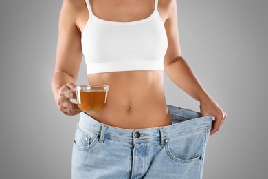 Young woman in old big jeans with cup of tea showing her diet results on grey background, closeup