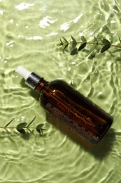 Bottle of hydrophilic oil and plant twigs in water on light green background, flat lay